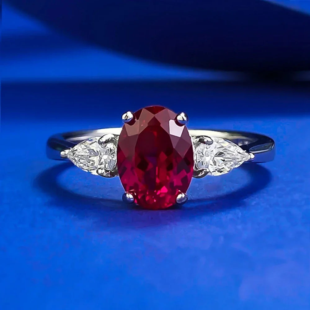 Lab-created ruby oval ring with pear-shaped moissanite side stones on silver band, blue background